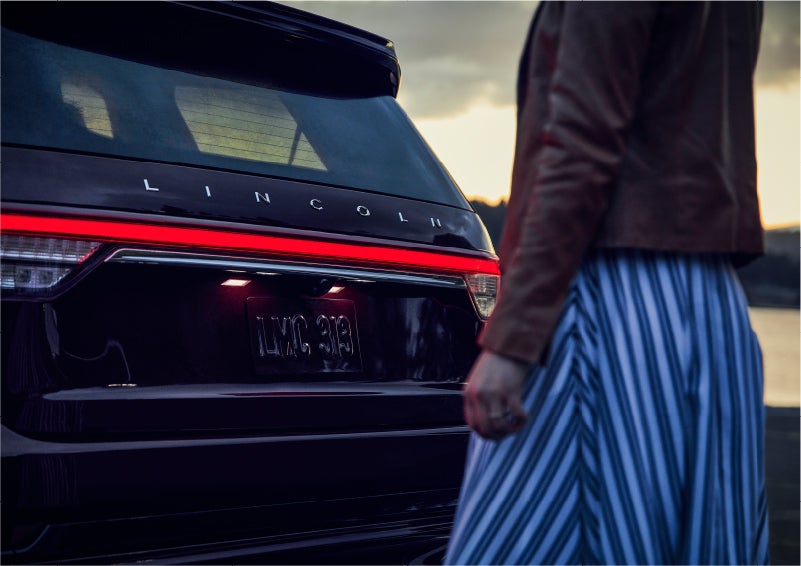 A person is shown near the rear of a 2023 Lincoln Aviator® SUV as the Lincoln Embrace illuminates the rear lights | Angela Krause Lincoln of Alpharetta in Alpharetta GA